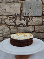 Small cake with white frosting on a white plate against a stone wall background, featuring a 'Happy Equinox' decoration.