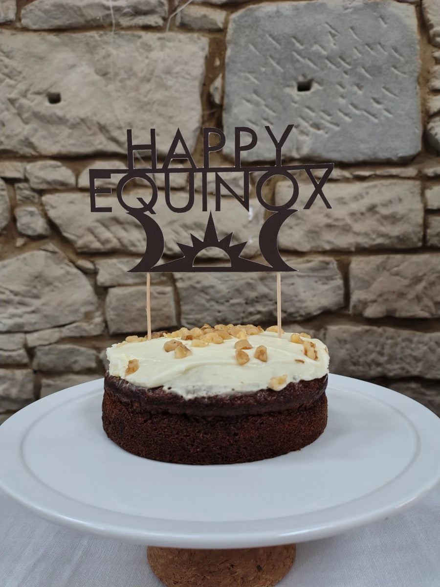 Small cake with white frosting on a white plate against a stone wall background, featuring a 'Happy Equinox' decoration.