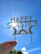 A holographic paper 'Happy Solstice' cake topper held by a hand against a bright blue sky with the sun shining through the center.