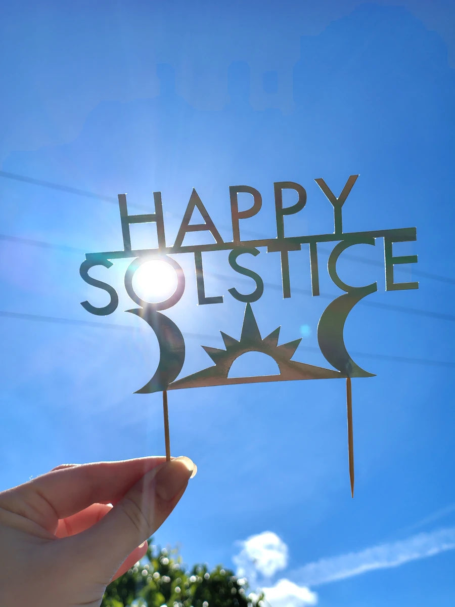 A holographic paper 'Happy Solstice' cake topper held by a hand against a bright blue sky with the sun shining through the center.