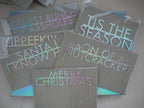 Six Christmas-themed cake toppers made with holographic card featuring various humorous and festive phrases. Sitting on a holographic box displayed on a light floor.