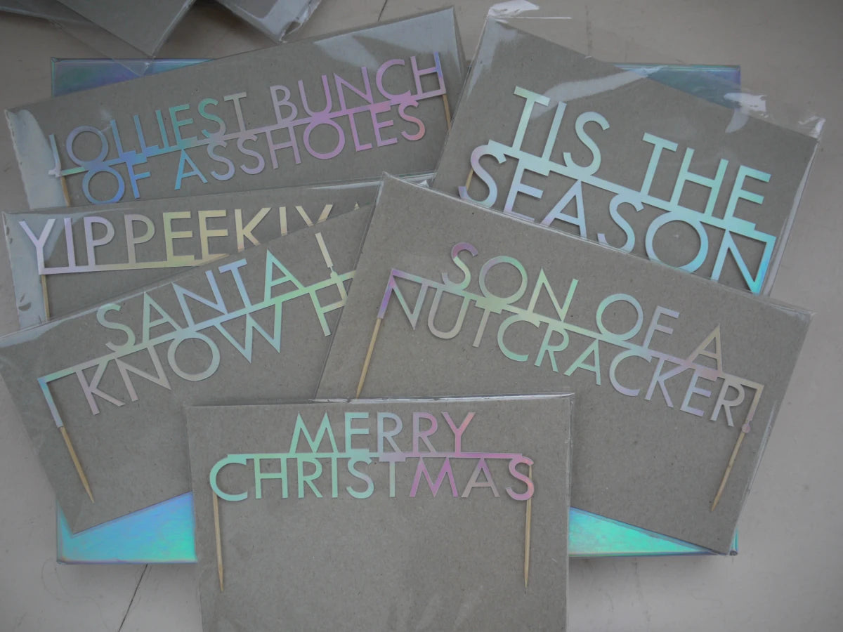 Six Christmas-themed cake toppers made with holographic card featuring various humorous and festive phrases. Sitting on a holographic box displayed on a light floor.
