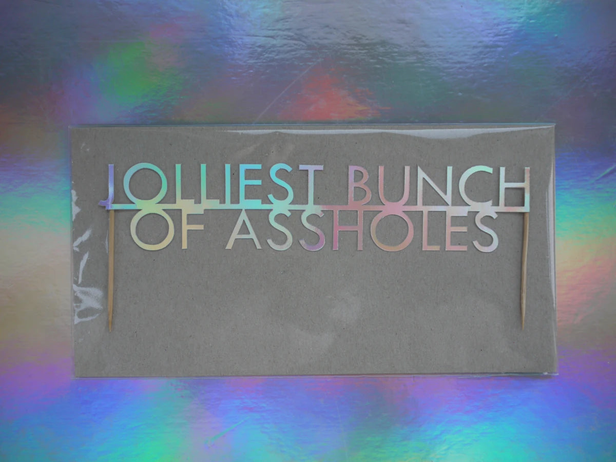 Christmas cake topper with the phrase 'JOLLIEST BUNCH OF ASSHOLES' in holographic card letters, set against a holographic, iridescent background.