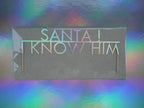 A Christmas cake topper that reads 'SANTA! I KNOW HIM' in holographic lettering, displayed against a colorful iridescent background.