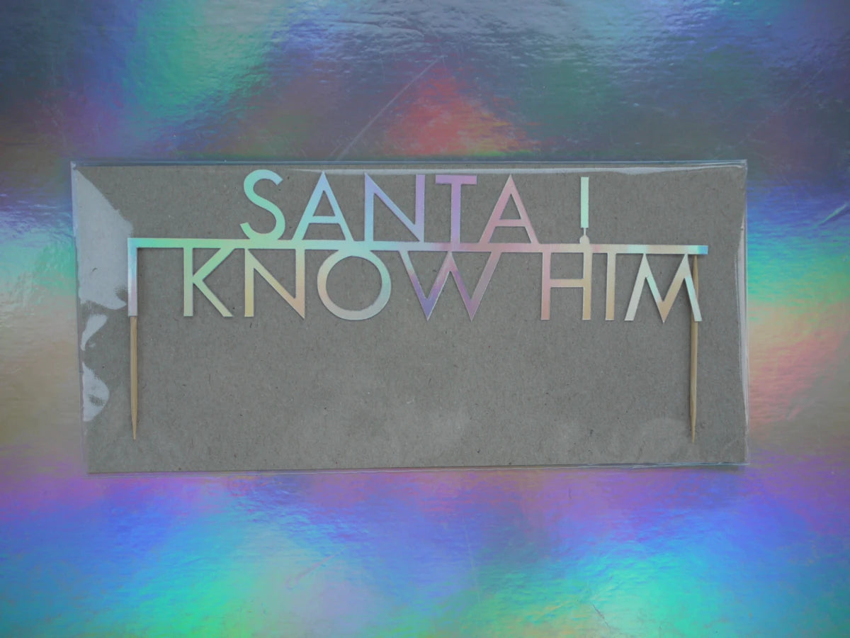 A Christmas cake topper that reads 'SANTA! I KNOW HIM' in holographic lettering, displayed against a colorful iridescent background.