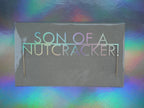 A 'SON OF A NUTCRACKER!' Christmas cake topper in holographic card. Displayed against a holographic, iridescent background.