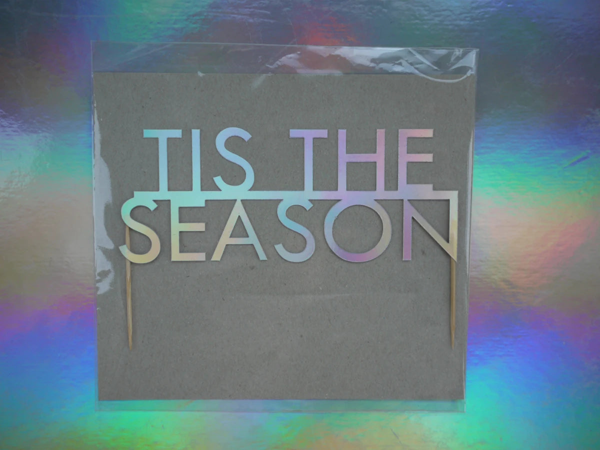 Holographic card sign reading 'TIS THE SEASON' as a fun festive Christmas cake topper. Photographed against a holographic rainbow background.