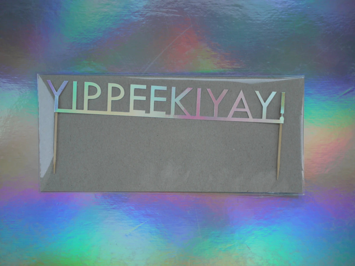 Holographic Christmas cake topper that reads “YIPPEEKIYAY!”, photographed against a holographic rainbow background.