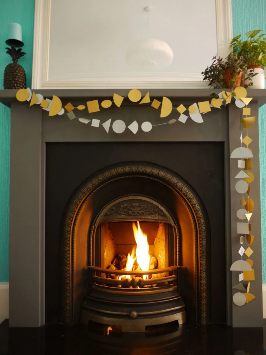 Decorative garlands made of gold and silver geometric shapes hangs across a fireplace mantel in a real interior setting, with a burning fireplace and surrounding room visible. Fun easy party themes to take away the stress of decorating