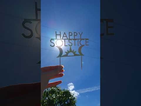 A holographic paper 'Happy Solstice' cake topper held by a hand against a bright blue sky with the sun shining through the center and birdsong heard throughout.