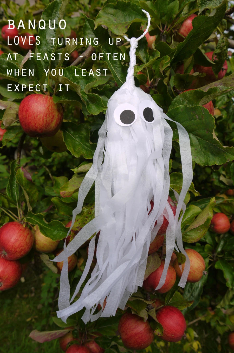 A white tissue paper ghost Halloween decoration with black eyes hangs from a tree branch with red apples and green leaves. White text on the left reads, 'BANQUO LOVES TURNING UP AT FEASTS - OFTEN WHEN YOU LEAST EXPECT IT'. Spooky but cute Halloween decoration.