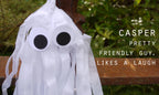 A white ghost-shaped hanging decoration with two large eyes and long white, photographed outdoors against a blurred green background with a brown post. White text on the right reads, 'CASPER PRETTY FRIENDLY GUY, LIKES A LAUGH'. Spooky but cute tassel Halloween decor.