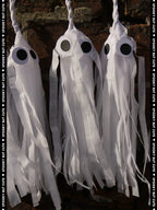 Three white paper tassel ghost Halloween decorations with large black eyes, shown against a dark rustic background with a vertical text watermark reading 'SPOOKY BUT CUTE'.
