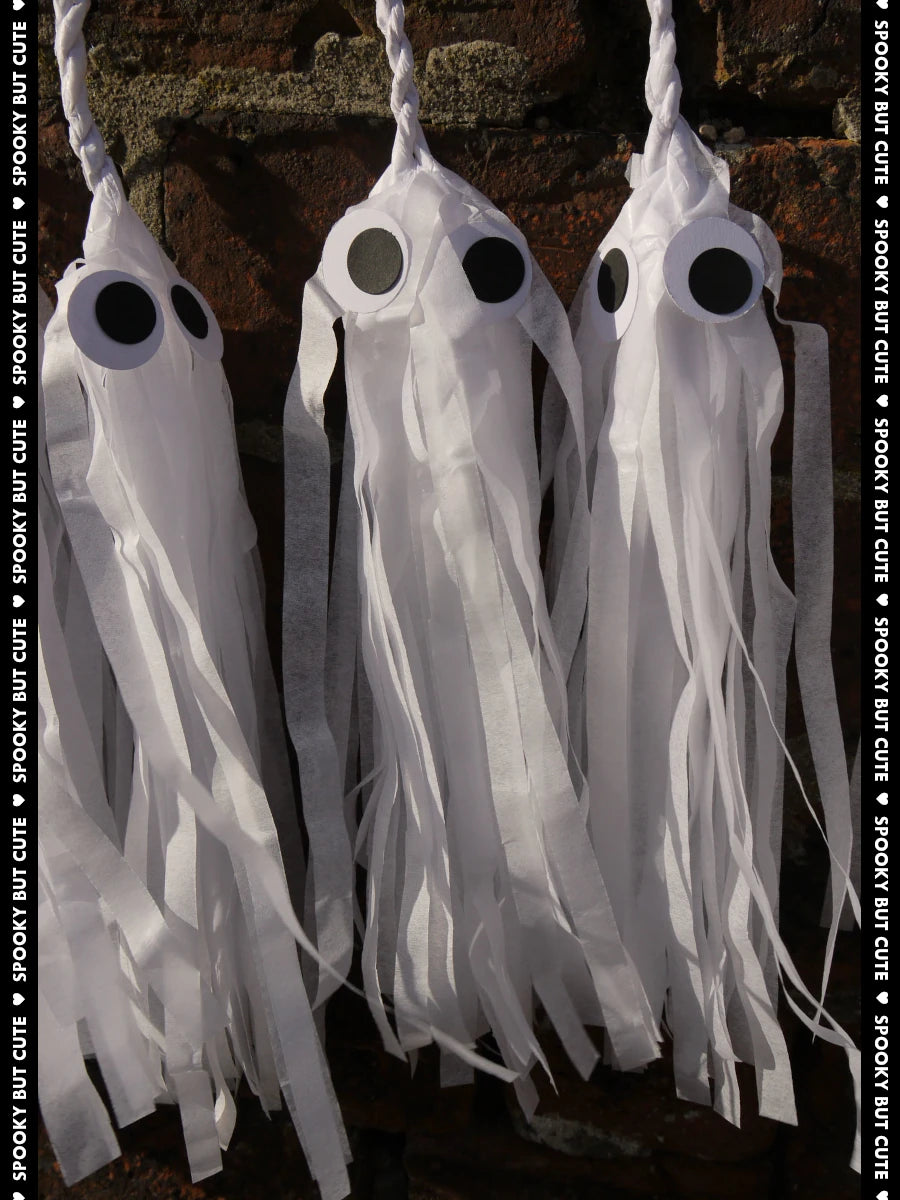Three white paper tassel ghost Halloween decorations with large black eyes, shown against a dark rustic background with a vertical text watermark reading 'SPOOKY BUT CUTE'.