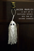 A white ghost-shaped tassel ornament with black eyes hangs from a gold owl door knocker on a dark brown wooden door. White text on the right reads 'JACOB MARLEY A BIT OF A RESTLESS SPIRIT, BIG FAN OF SECOND CHANCES'. Spooky but cute Halloween decor.