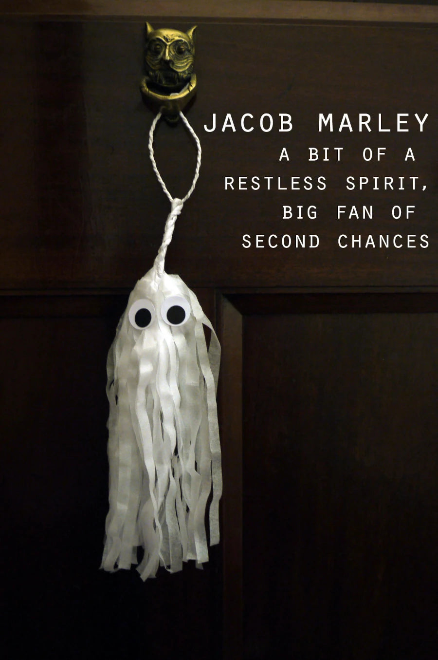 A white ghost-shaped tassel ornament with black eyes hangs from a gold owl door knocker on a dark brown wooden door. White text on the right reads 'JACOB MARLEY A BIT OF A RESTLESS SPIRIT, BIG FAN OF SECOND CHANCES'. Spooky but cute Halloween decor.
