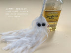 A handmade tissue paper tassel ghost decoration, shown next to a bottle of Jose Cuervo Especial tequila on a light beige surface. Text on the left reads 'JIMMY ANGELOV LIKES ELVIS, TEQUILA AND JELLY DOUGHNUTS - WITH CREAM'. Spooky but cute Halloween decor.