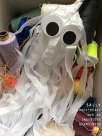 A handmade ghost decoration made from white tissue paper with spooky but cute eyes, nestled inside a box surrounded by colorful spools of thread. A watermark in the bottom right corner reads 'SALLY SWEETHEART AND AN INGENIOUS SEAMSTRESS'.